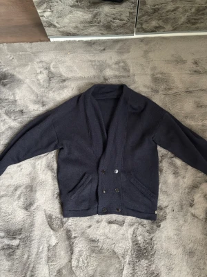 Cardigan - Mysig mörkblå stickad kofta med sjalkrage och dubbelknäppning framtill. Koftan har två fickor och ribbade muddar vid ärmslut och nederkant. Perfekt för lager på lager och en avslappnad stil.