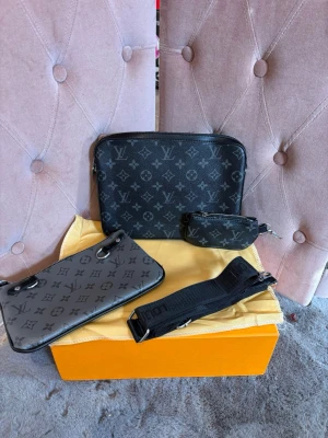 Louis Vuitton axelväska monogram svart - Snygg svart axelväska från Louis Vuitton med klassiskt monogrammönster i grått. Väskan har flera delar, inklusive en mindre pouch och justerbar axelrem i textil. Tillverkad i slitstarkt läder med silverfärgade detaljer. Perfekt för dig som gillar exklusiva accessoarer.