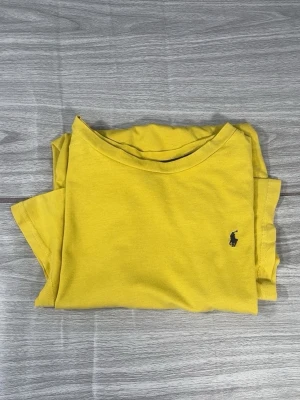 Gul t-shirt från Polo Ralph Lauren - Snygg gul t-shirt från Polo Ralph Lauren med klassisk blå logga broderad på bröstet. T-shirten har rund halsringning och korta ärmar. Tillverkad i mjuk bomull som känns skön mot huden. Perfekt för dig som vill ha en enkel men stilren look.