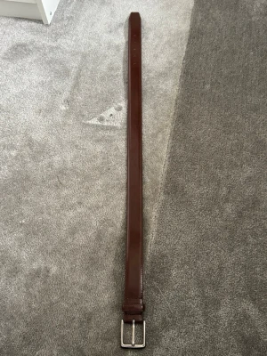 Brunt skärp i skinn med silverfärgat spänne - Stilrent brunt bälte i skinn med klassiskt silverfärgat spänne. Perfekt accessoar för att lyfta din outfit och passar till både jeans och kostymbyxor. Enkel design utan mönster och med flera hål för justerbar passform.