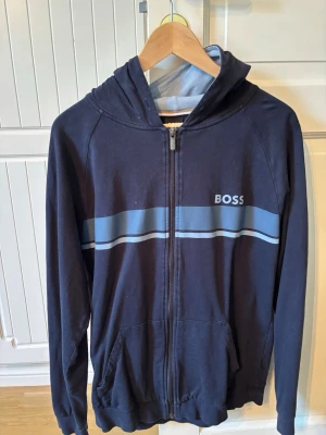 Marinblå hoodie från BOSS - Snygg marinblå hoodie från BOSS med dragkedja framtill och ljusblåa ränder över bröstet. Hoodien har fickor på sidorna och en huva med ljusblå insida. Perfekt för en avslappnad och stilren look.
