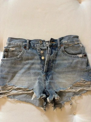 Jeansshorts från Zara - Säljer ett par ljusblå jeansshorts från Zara med slitna detaljer och råa kanter. Perfekta till sommaren i storlek 32.