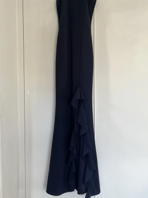 Marinblå långklänning med volang - Elegant marinblå långklänning med figurnära passform och volangdetaljer längs nederdelen. Klänningen är ärmlös och har en stilren, enkel design som ger ett exklusivt intryck. Perfekt för bal.