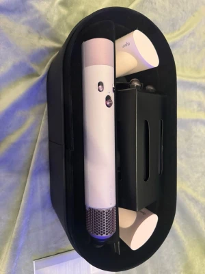 Dyson hårstyler med tillbehör - Säljer en vit Dyson hårstyler med flera utbytbara tillbehör, perfekt för att skapa olika frisyrer. Kommer i en svart förvaringsbox och inkluderar borsthuvuden och stylingmunstycken. Modern design och högkvalitativt material. 