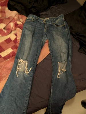 Blå slitna jeans med hål på knäna - Säljer ett par blå jeans med tydliga slitningar och stora hål på båda knäna. Klassisk femficksmodell med knapp och dragkedja. Jeansen har en rak passform och är tillverkade i denim. Perfekta för en avslappnad och trendig look.