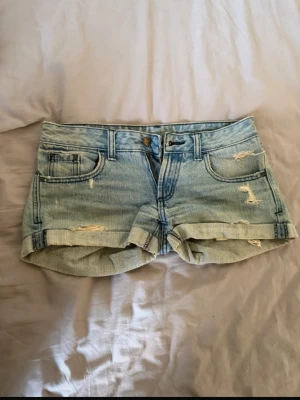 Ljusblå denimshorts med slitningar - Säljer ett par ljusblå denimshorts från Never Den med slitna detaljer och upprullade benslut. Klassisk femficksmodell med bälteshällor och knappgylf. Perfekta för sommaren och ger en avslappnad look. Lågmidjande!
