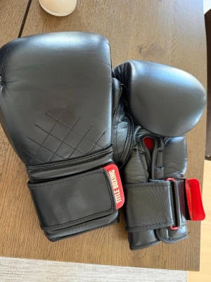 Svarta boxningshandskar Title Boxing - Säljer ett par svarta boxningshandskar från Title Boxing, 16oz. Handskarna har bred kardborrestängning och detaljer i rött. Tillverkade i slitstarkt konstläder med vadderad ovansida för extra skydd. Perfekta för träning eller sparring. Handskarna är använda 3 gånger. 