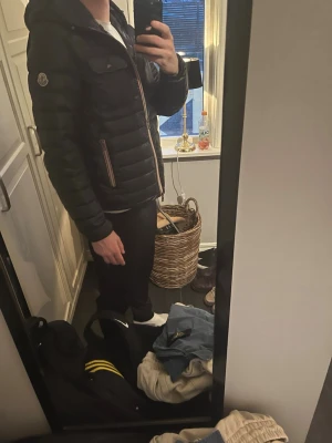Svart dunjacka från Moncler med huva - Snygg svart dunjacka från Moncler med huva och quiltad design. Jackan har dragkedja framtill, två fickor med dragkedja och en Moncler-logga på ärmen. Perfekt för vår, höst och vinter och riktigt stilren look.