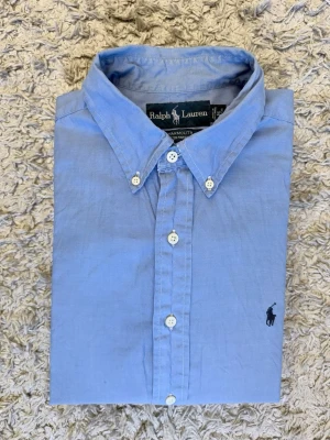 Blå skjorta från Ralph Lauren - Klassisk ljusblå skjorta från Ralph Lauren med button-down krage och broderad logga på bröstet. Skjortan har långa ärmar och vita knappar framtill. Perfekt för en stilren och avslappnad look.