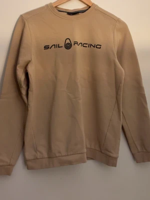 Beige sweatshirt från Sail Racing - Stilren beige sweatshirt från Sail Racing med svart logotyptryck på bröstet. Tröjan har rund halsringning, långa ärmar och ribbade muddar vid ärmslut och nederkant. Perfekt för en avslappnad och trendig look.