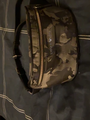 Kamouflagemönstrad magväska Armani - Snygg magväska från Emporio Armani i kamouflagemönstrat tyg med svarta och grå toner. Väskan har justerbart band, två dragkedjefack och logga framtill. Perfekt för att bära det viktigaste på ett stilrent sätt.