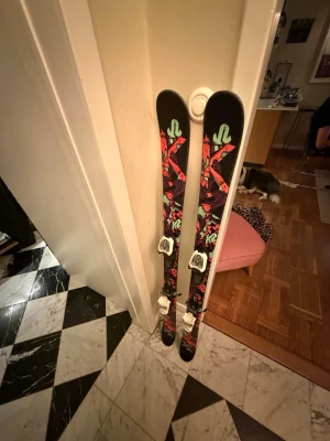 Svarta K2 twintip skidor med bindningar - Säljer ett par svarta twintip skidor från K2 med färgglad grafik i rött, grönt och lila. Skidorna har vita bindningar och är perfekta för dig som gillar park eller all mountain. Grym design som sticker ut i backen! Måste bli avhämtade.