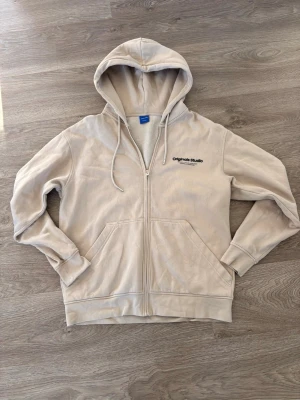 Beige zip hoodie Originals Studio - Snygg beige hoodie med dragkedja från Originals Studio. Klassisk modell med huva, dragsnören och två stora fickor framtill. Broderad logga på bröstet. Tillverkad i mjukt bomullsmaterial som känns skönt mot huden. Perfekt för en avslappnad och stilren look.