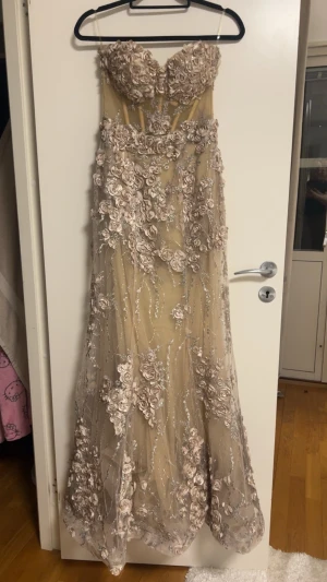 Champagnefärgad långklänning med spets - Elegant långklänning i champagnefärg med detaljerade spetsapplikationer och 3D-blommor över hela klänningen. Klänningen har en romantisk look med transparent lager och skimrande inslag. Perfekt för bal eller festliga tillfällen.
