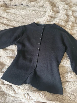 Svart kofta från Gina Tricot - Stilren svart kofta från Gina Tricot med rund halsringning och knappar längs hela framsidan. Koftan har en rak passform och är tillverkad i ett mjukt, tunt stickat material som passar perfekt till lager på lager. Enkel att matcha med olika outfits.