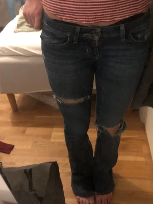 Mörkblå slitna bootcut jeans - Säljer ett par mörkblå bootcut jeans med slitningar och hål vid knäna. Jeansen har klassisk femficksmodell och låg midja. Perfekta för en avslappnad och trendig look. Materialet är denim i bomull. LÅGMIDJADE 