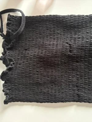 Svart bandeau tubtopp från XOXO - Säljer en svart bandeau tubtopp från XOXO med smockad struktur och elastiska kanter. Toppen är ärmlös och har en enkel, stilren look som passar perfekt till många outfits. Materialet känns mjukt och stretchigt.