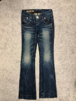 Blå flare jeans från Red Pepper - midja rakt över:34. lår: 23. inre ben: 84. Säljer ett par blå bootcut jeans från Red Pepper med slitningar och broderade detaljer på bakfickorna. Jeansen har låg midja, dekorativa knappar och kontrastsömmar. Materialet är klassisk denim i bomull med stretch för extra komfort.