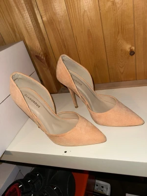 Persikofärgade pumps från Glamorous - Snygga persikofärgade pumps från Glamorous med spetsig tå och hög smal klack. Skorna är i mockaimitation och har en öppen sida som ger en trendig look. Perfekta för att lyfta vilken outfit som helst.