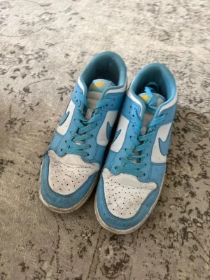 Nike Dunk Low blå och vita sneakers - Snygga Nike Dunk Low sneakers i blått och vitt med klassisk swoosh på sidan. Skorna har perforerad tåbox, vita paneler och blå detaljer samt matchande blå skosnören. Ovandelen är i läder och innerfodret är mjukt för extra komfort.