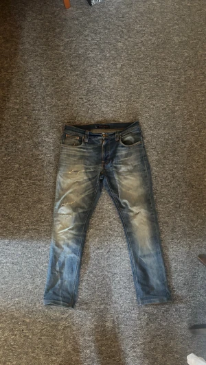 Nudie  - Säljer ett par blå jeans från Nudie Jeans med slitningar och tvättade detaljer på benen. Det är slim passform och modellen heter thin finn. Byxorna ör i hyfsat bra skick men det är hål i fickan. Jag kunde ha byxorna ändå och mobilen trillar inte ut om man lägger i den rätt. Annars är det inte svårt att fixa. Pris diskuterbart