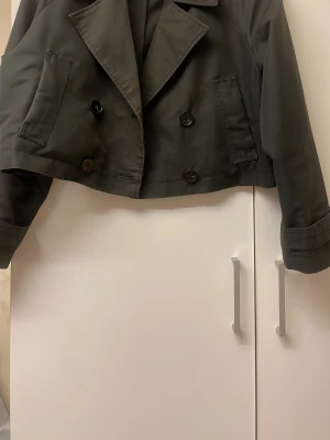 Kort svart trenchcoat med knappar - Snygg kort trenchcoat i svart med dubbelknäppning framtill och bred krage. Jackan har två snedställda fickor och dekorativa knappar vid ärmsluten. Perfekt för dig som gillar en stilren och trendig look.    SVARTA FÄRGEN HAR ÄNDRATS PÅ VISSA STÄLLEN PGA SOLEN! 
