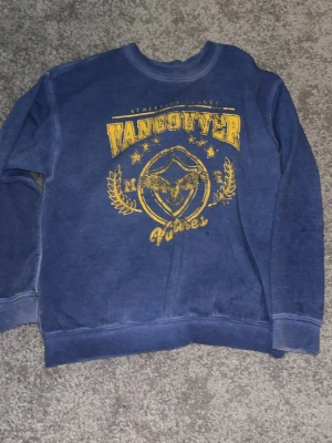 Blå hoodie med Vancouver-tryck - Cool blå hoodie med gult Vancouver-tryck och grafiskt motiv framtill. Rund halsringning och ribbade muddar vid ärmar och nederkant. Perfekt för dig som gillar streetwear och sportig stil.
