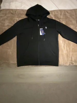  Polo Ralph Lauren Kofta - Snygg svart hoodie från Polo Ralph Lauren med dragkedja framtill och klassisk logga broderad på bröstet. Hoodien har huva med dragsko och två fickor framtill. Tillverkad i mjukt material som känns skönt mot huden. Perfekt för en avslappnad och stilren look.