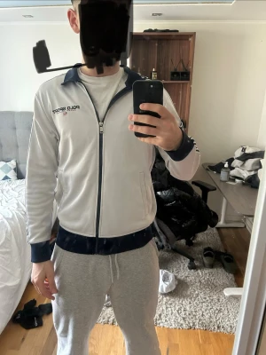 Vit track jacket från Polo Sport - Säljer en vit track jacket från Polo Sport med marinblå detaljer på krage, muddar och nederkant. Dragkedja framtill och logga på bröstet samt text längs ärmarna. Perfekt för en sportig och avslappnad stil.