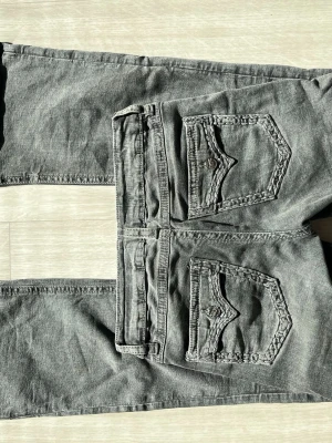 Grå jeans från ginatricot med fickdetaljer - Säljer ett par grå jeans från ginatricot med raka ben och två stora fickor med lock och knapp bak. Jeansen har en cool tvättad look och tydliga sömmar som ger extra edge. Perfekta för dig som gillar streetstyle och vill ha något unikt i garderoben.