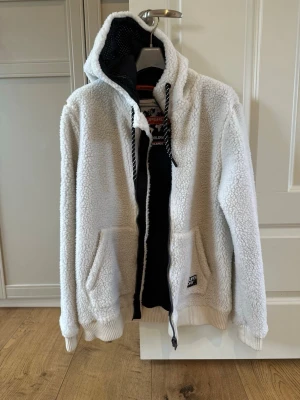 Vit teddyjacka från Superdry - Mysig vit teddyjacka från Superdry med huva och dragkedja. Jackan har två stora fickor framtill, ribbade muddar och svart innerfoder. Detaljer med snörning i huvan och Superdry-logga på ena fickan. Perfekt för kalla dagar. Endast testad 