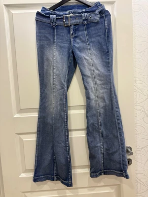 Blå bootcut jeans med bälte - Säljer ett par blå bootcut jeans med markerade sömmar framtill och ett brett, matchande bälte i midjan. Jeansen har en lätt tvättad look och utsvängda ben för en retro vibe. Perfekta för dig som gillar en trendig och avslappnad stil.