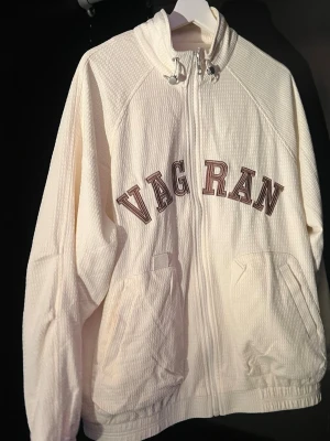 Vit track jacket från VAG RAN - Vit track jacket i strukturerat tyg med dragkedja framtill och två fickor. Bruna bokstäver 'VAG RAN' på bröstet och tre bruna ränder längs ena ärmen. Liten patch med texten 'Vuitton' på ryggen. Sportig och avslappnad stil, perfekt för streetwear.