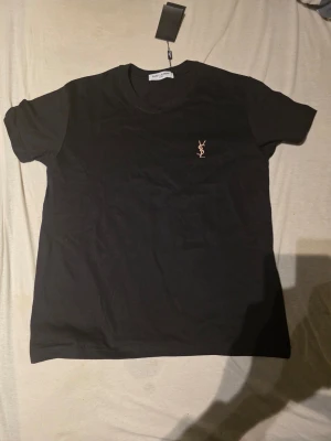 Svart t-shirt från Saint Laurent - Snygg svart t-shirt från Saint Laurent med klassisk rund hals och korta ärmar. Diskret YSL-logga broderad i guld på bröstet. Tillverkad i mjuk bomull för en bekväm känsla och stilren look.