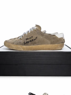 Saint Laurent  - Säljer ett par beigea Saint Laurent sneakers i mocka med vit sula och vita skosnören. Bara skorna. Större skador finns i bild 4 och 5 finns hål.  Sidan har handskriven logga och skorna har en klassisk, låg profil med rund tå. Perfekta för dig som gillar stilrena och exklusiva sneakers. 