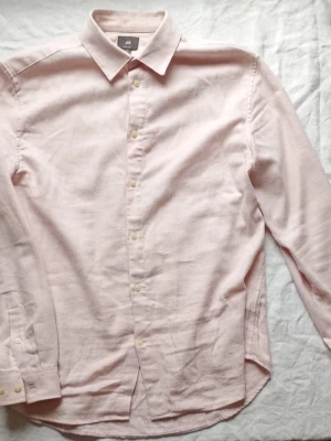 Ljusrosa skjorta från H&M - Säljer en stilren ljusrosa skjorta från H&M med klassisk krage och knappar framtill. Skjortan är långärmad och tillverkad i mjuk bomull, perfekt för en clean och enkel look.