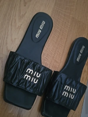 Svarta sandaler  - Stilrena svarta sandaler med breda, quiltade remmar och logotyp i silver framtill. Sandalerna har fyrkantig tå och är tillverkade i ett glansigt material som ger en lyxig känsla. Perfekta för en trendig och avslappnad look.