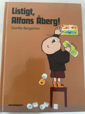 Listigt, Alfons Åberg! - En charmig barnbok med hård pärm, skriven av Gunilla Bergström. Omslaget är brunt med en illustration av Alfons Åberg som står på en stol och försöker nå kakor. Perfekt för dig som gillar klassiska svenska berättelser.