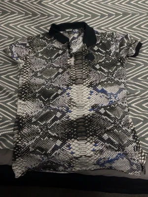 Ormmönstrad pikétröja med svart krage - Säljer en unik pikétröja med ormmönster i svart, vit och grå med inslag av blått. Tröjan har korta ärmar, svart krage och knappar framtill. Perfekt för dig som vill sticka ut med ett djuriskt mönster.