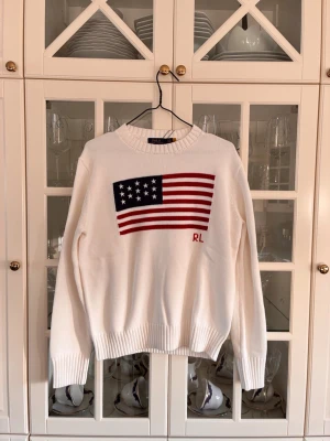 Vit stickad tröja Polo Ralph Lauren - Vit stickad tröja från Polo Ralph Lauren med amerikansk flagga i rött, vitt och blått på bröstet samt RL-broderi. Tröjan har rund halsringning och ribbade muddar vid ärmslut och nederkant. Perfekt för en klassisk och avslappnad stil.