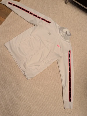 Unik Nike tröja - Säljer en vit träningsjacka från Nike med hel dragkedja och hög krage. Jackan har röda och svarta detaljer längs ärmarna med Nike-loggor, samt en röd Nike Swoosh på bröstet. Perfekt för sport eller chill. Materialet är mjukt och stretchigt, troligen polyester.