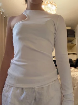 Vit oneshoulder topp med cut out - Trendig vit topp med oneshoulder-design och unik cut out-detalj vid halsen. Toppen är tight och har en lång ärm på ena sidan medan den andra axeln är bar. 