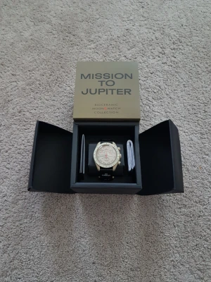 Omega x Swatch Mission to Jupiter - Säljer en Omega x Swatch Mission to Jupiter från Bioceramic MoonSwatch-kollektionen. Klockan har en beige urtavla med svarta och röda detaljer, svart band och klassisk kronografdesign. Perfekt för dig som gillar unika och ikoniska klockor.