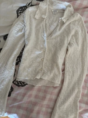 Vit strukturerad blus från Monki - Säljer en vit blus från Monki med strukturerat tyg och krage. Blusen har långa ärmar och knäppning framtill med små vita knappar. Materialet är lätt och ger en cool textur. Perfekt för dig som gillar en unik och trendig look.