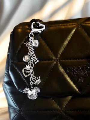 Silverfärgad väsksmycke med berlocker - Snyggt silverfärgat väsksmycke med flera berlocker, inklusive ett glittrigt musmotiv, hjärta, krona och texten 'Princess'. Perfekt att hänga på väskan för en personlig touch. Smycket har en hjärtformad karbinhake och är dekorerat med gnistrande stenar.
