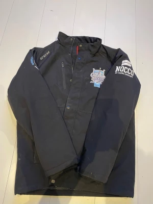 Svart hockeyjacka från CCM x NOCCO - Svart jacka från CCM med Outdoor Hockey Stockholm- och NOCCO-tryck på bröst och ärm. Jackan har hög krage, dragkedja och knappar framtill samt en bröstficka med dragkedja. Perfekt för dig som gillar hockey och sportig stil.