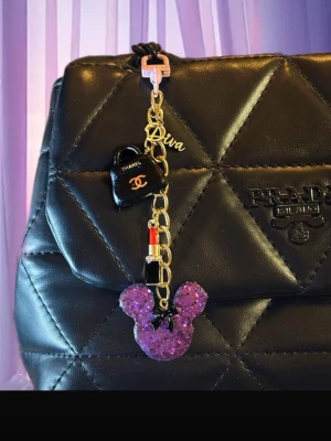 Väsksmycke/nyckelring med charms - Nyckelring med lila glittrigt musöron-hänge, svart väska, röd läppstift-charm och texten 'Diva'. Kedjan är guldfärgad och detaljerna är i plast och metall. Perfekt som accessoar till väskan eller nycklarna.