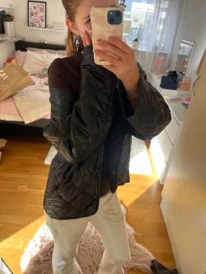 Svart quiltad vindjacka - Säljer en svart vindjacka med quiltat mönster och tryckknappar framtill. Jackan har en rak passform och är tillverkad i ett glansigt syntetmaterial som är lätt och smidigt. Perfekt för lager-på-lager och enkel att matcha med olika outfits.