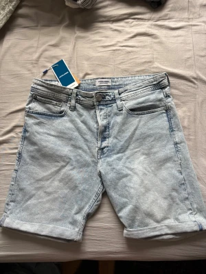 Ljusblå denimshorts från Jack & Jones - Snygga ljusblå denimshorts från Jack & Jones med klassisk femficksdesign och uppvikta benslut. Shortsen har bälteshällor och knappgylf. Perfekta för varma dagar och enkel att matcha med olika stilar.