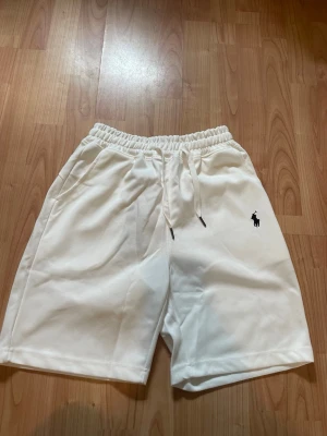 Vita shorts från Polo Ralph Lauren - Snygga vita shorts från Polo Ralph Lauren med elastisk midja och snörning. Klassisk logga broderad i svart på vänster ben. Tillverkade i mjuk bomull för skön känsla. Perfekta för sommardagar och chill häng.